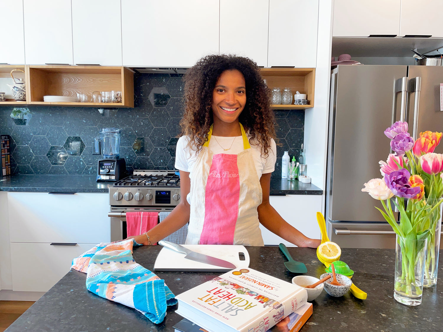 Home cook Alia Marie standing in her kitchen smiling at the camera. In front of her on the island are cookbooks, knives and cutting boards, and vases of flowers.